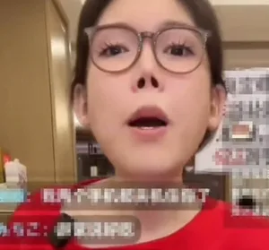 浪胃仙在直播画面中正面出镜,佩戴黑框眼镜,鼻部明显不对称,左侧鼻孔较大,表情平静直视镜头,背景为简易室内直播环境。