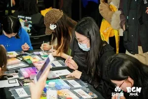 新物集BGM游戏展上海站现场照片:多人围站在一个摆放着多款潮玩手办和解压捏捏玩具的展台前观看或挑选,背景可见展会标识与灯光装饰。